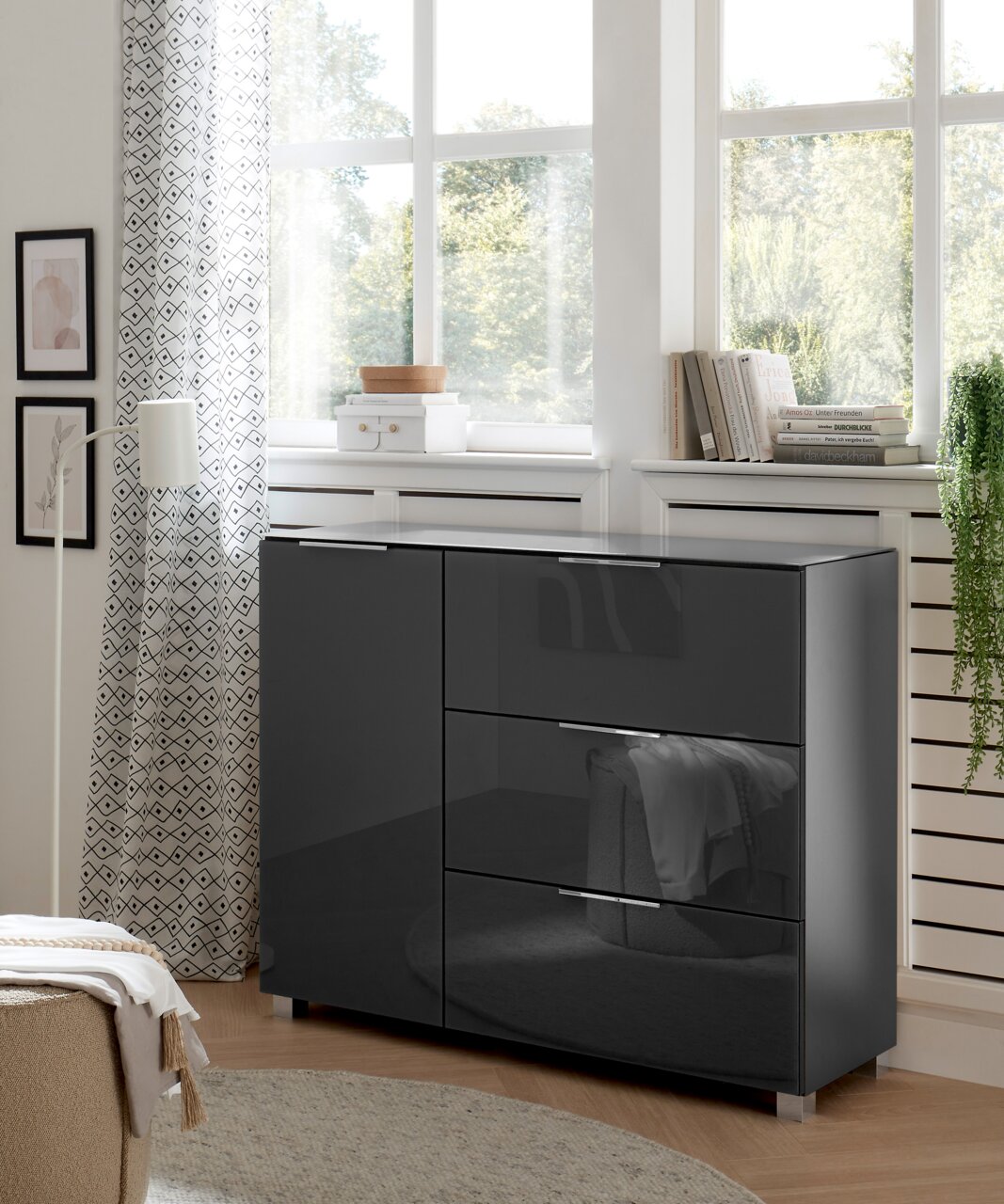 XORA Kommode LIPPO Moderne, dunkelgraue Kommode mit Hochglanzoberfläche, seitliche Perspektive, in einem hellen Raum mit großen Fenstern und dekorativen Elementen.