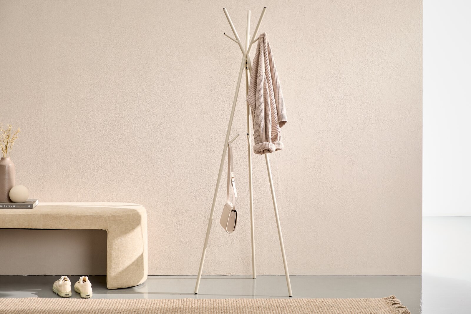 Freistehende weiße Garderobenständer/Standgarderobe mit mehreren Haken, daran ein beiger Mantel und eine kleine Tasche, im minimalistischen Flur neben einer gepolsterten Bank Frontalansicht auf Augenhöhe.