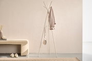 Freistehende weiße Garderobenständer/Standgarderobe mit mehreren Haken, daran ein beiger Mantel und eine kleine Tasche, im minimalistischen Flur neben einer gepolsterten Bank; Frontalansicht auf Augenhöhe.