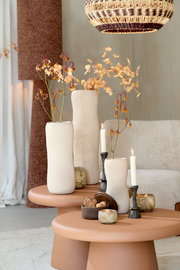 Drei elegante, cremefarbene Vasen mit Blumenarrangements auf einem runden, terrakottafarbenen Tisch, umgeben von Kerzen und dekorativen Elementen, aus einer seitlichen Perspektive betrachtet.