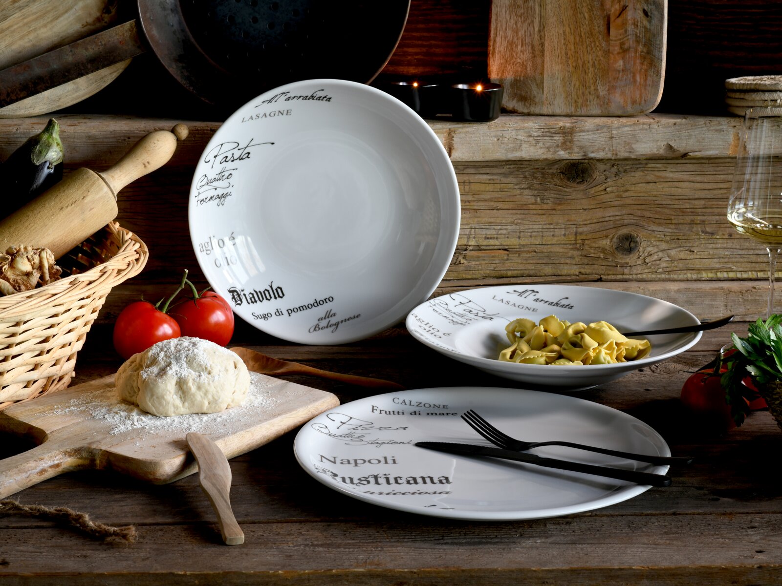 Rustikales Stillleben mit weißem Porzellan-Geschirrset mit italienischer Schrift, großer Schüssel und zwei Tellern (einer mit Pasta), auf Holztisch mit Teig, Tomaten und Küchenutensilien, fotografiert in schräger Frontansicht.