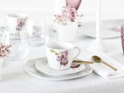 Weiße Porzellantasse mit rosa Orchideenmotiv auf passender Untertasse und Teller, neben goldenem Löffel auf weißer Serviette, fotografiert in schräger Frontansicht auf gedecktem Tisch.