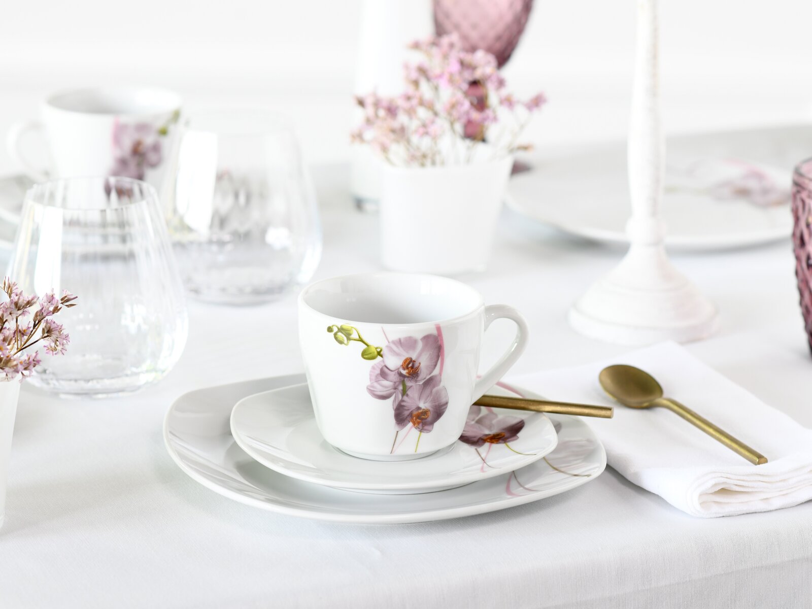 Weiße Porzellantasse mit rosa Orchideenmotiv auf passender Untertasse und Teller, neben goldenem Löffel auf weißer Serviette, fotografiert in schräger Frontansicht auf gedecktem Tisch.