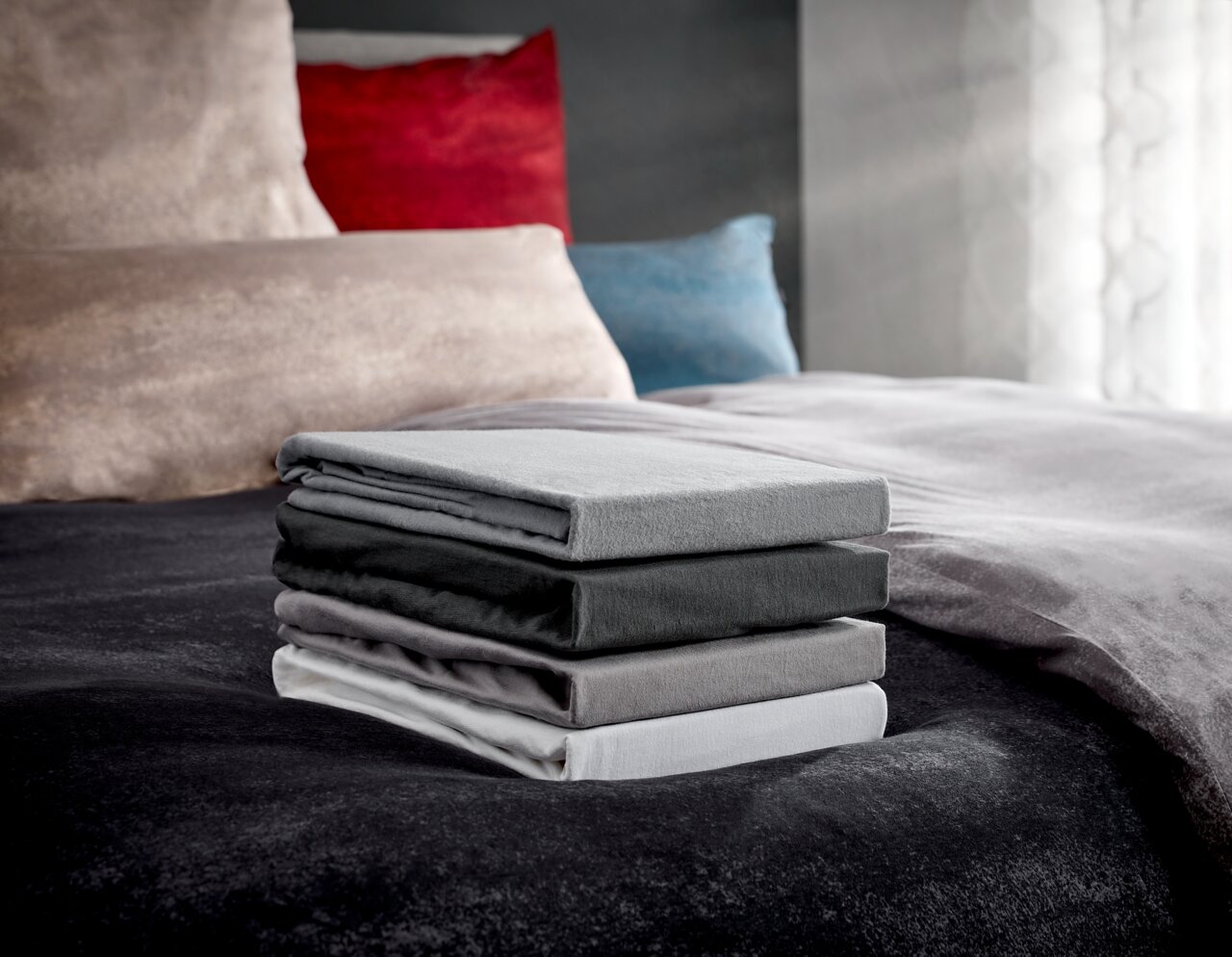 Novel Topper-Spannbetttuch Florenz Ein Stapel von vier gefalteten Spannbetttüchern in verschiedenen Grautönen auf einem Bett, fotografiert aus einer seitlichen Perspektive.