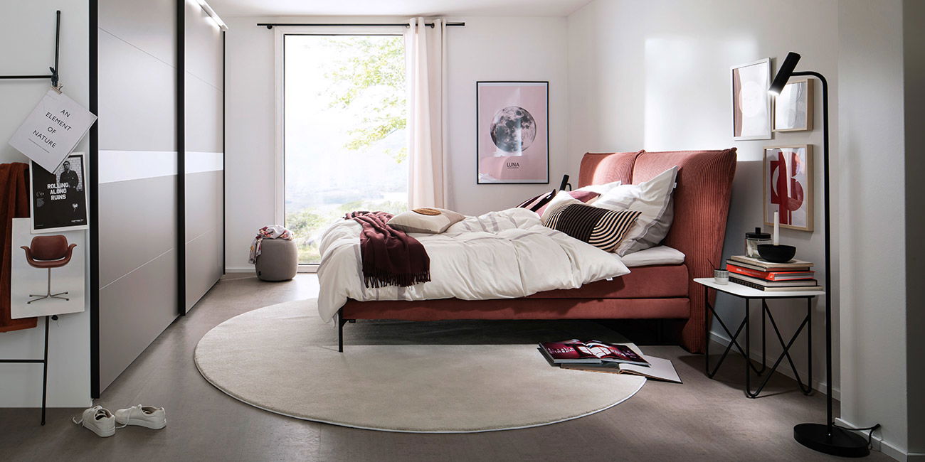 Helles Schlafzimmer mit rosa Bett, weißer Bettwäsche, Deko-Bildern und großem Fenster.