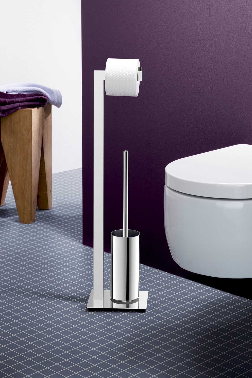 Freistehende WC-Garnitur aus poliertem Edelstahl mit Toilettenpapierhalter und Toilettenbürste neben einem weißen WC vor violetter Wand, Frontansicht in leicht schräger Perspektive von links.