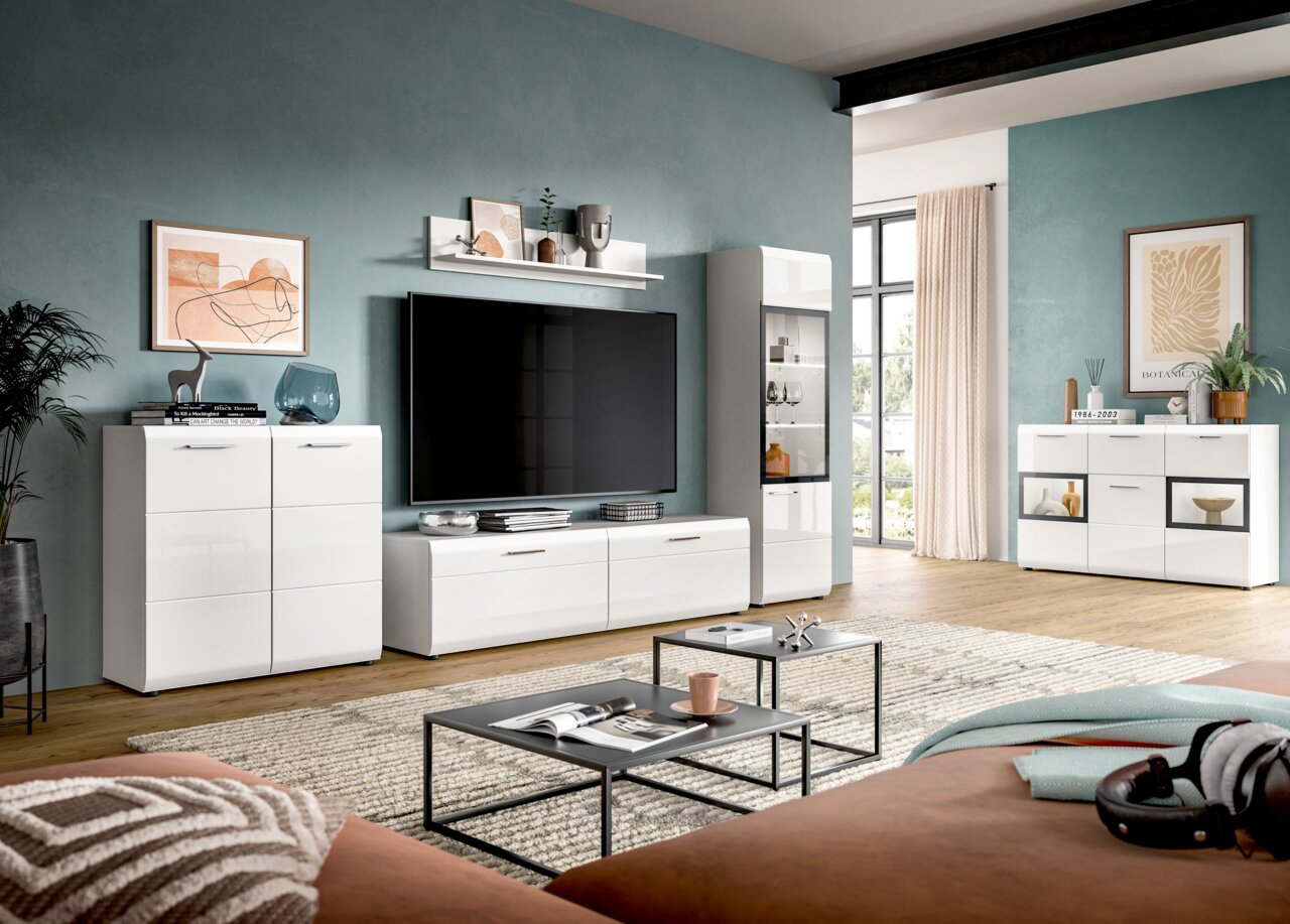 Modernes Wohnzimmer-Set in Weiß mit TV-Lowboard, Vitrine und Sideboards vor petrolfarbener Wand, frontal aus leicht seitlicher Perspektive aufgenommen.