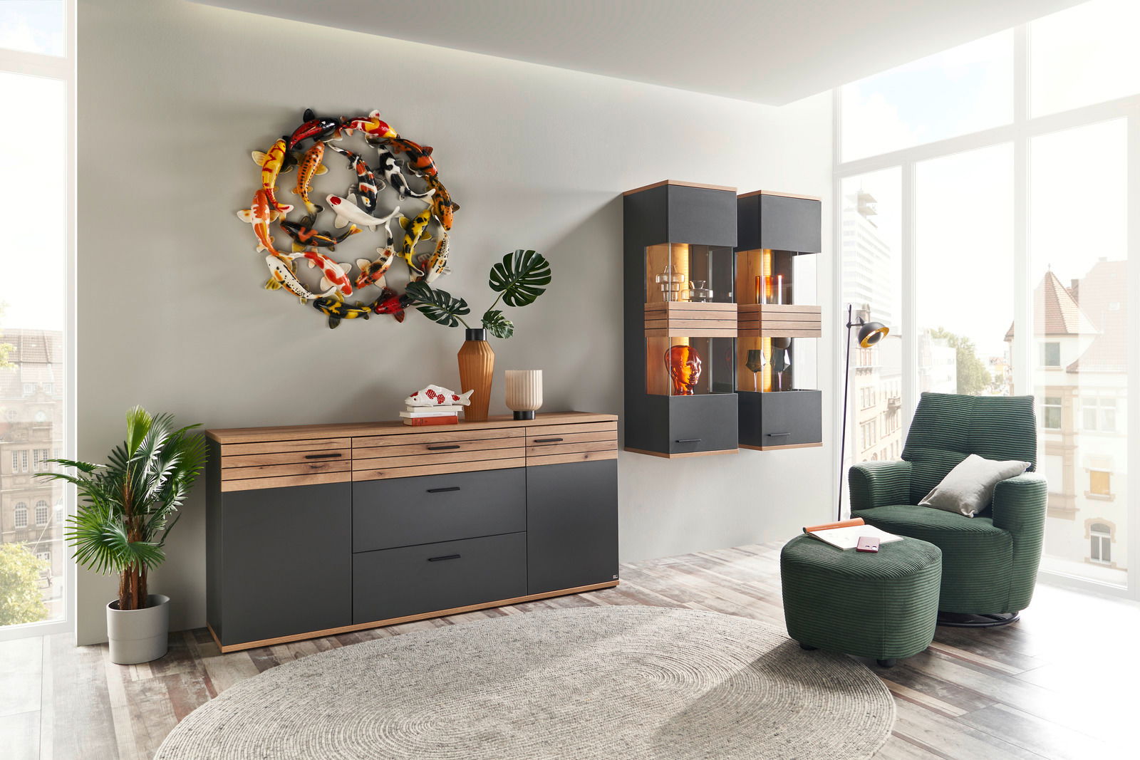 Moderne Wohnzimmeransicht mit einem stilvollen Sideboard in Holzoptik und dunklem Finish, dekoriert mit Pflanzen und Kunstobjekten, sowie einer gemütlichen Sitzecke mit grünem Sessel und Hocker. Perspektive von vorne.