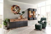 Moderne Wohnzimmeransicht mit einem stilvollen Sideboard in Holzoptik und dunklem Finish, dekoriert mit Pflanzen und Kunstobjekten, sowie einer gemütlichen Sitzecke mit grünem Sessel und Hocker. Perspektive von vorne.