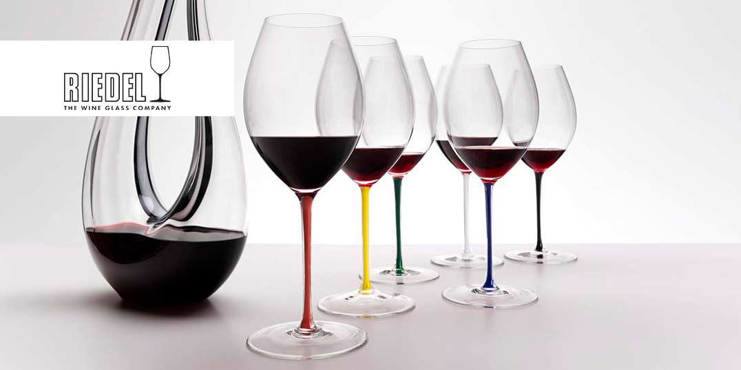 Sechs Weingläser mit bunten Stielen und eine Karaffe mit Rotwein auf einem weißen Tisch.