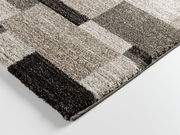 Nahaufnahme eines rechteckigen Teppichs mit geometrischem Muster in verschiedenen Grautönen, perspektivisch von der Seite aufgenommen.