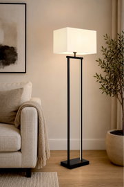 Moderne schwarze Stehlampe mit quadratischem weißem Stoffschirm, eingeschaltet neben einem beigen Sofa im Wohnzimmer; Perspektive: frontale, leicht seitliche Ansicht in Raumumgebung.