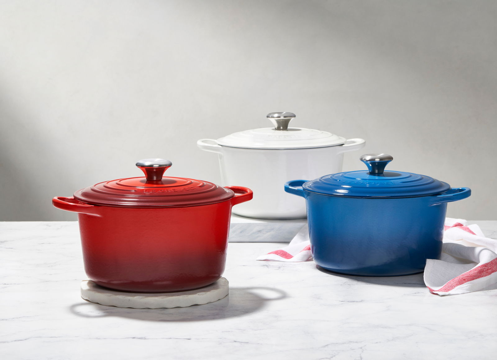 Drei runde Bräter in Rot, Weiß und Blau mit Deckeln und Griffen, auf einer Marmoroberfläche von vorne fotografiert.