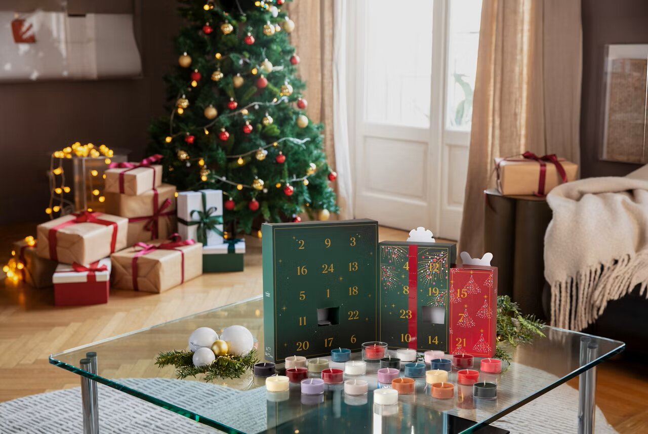 Luxe adventskalender met 24 kleurrijke geurkaarsjes, van voren in driekwartperspectief gepresenteerd op een glazen tafel in een sfeervol versierde kerstwoonkamer.