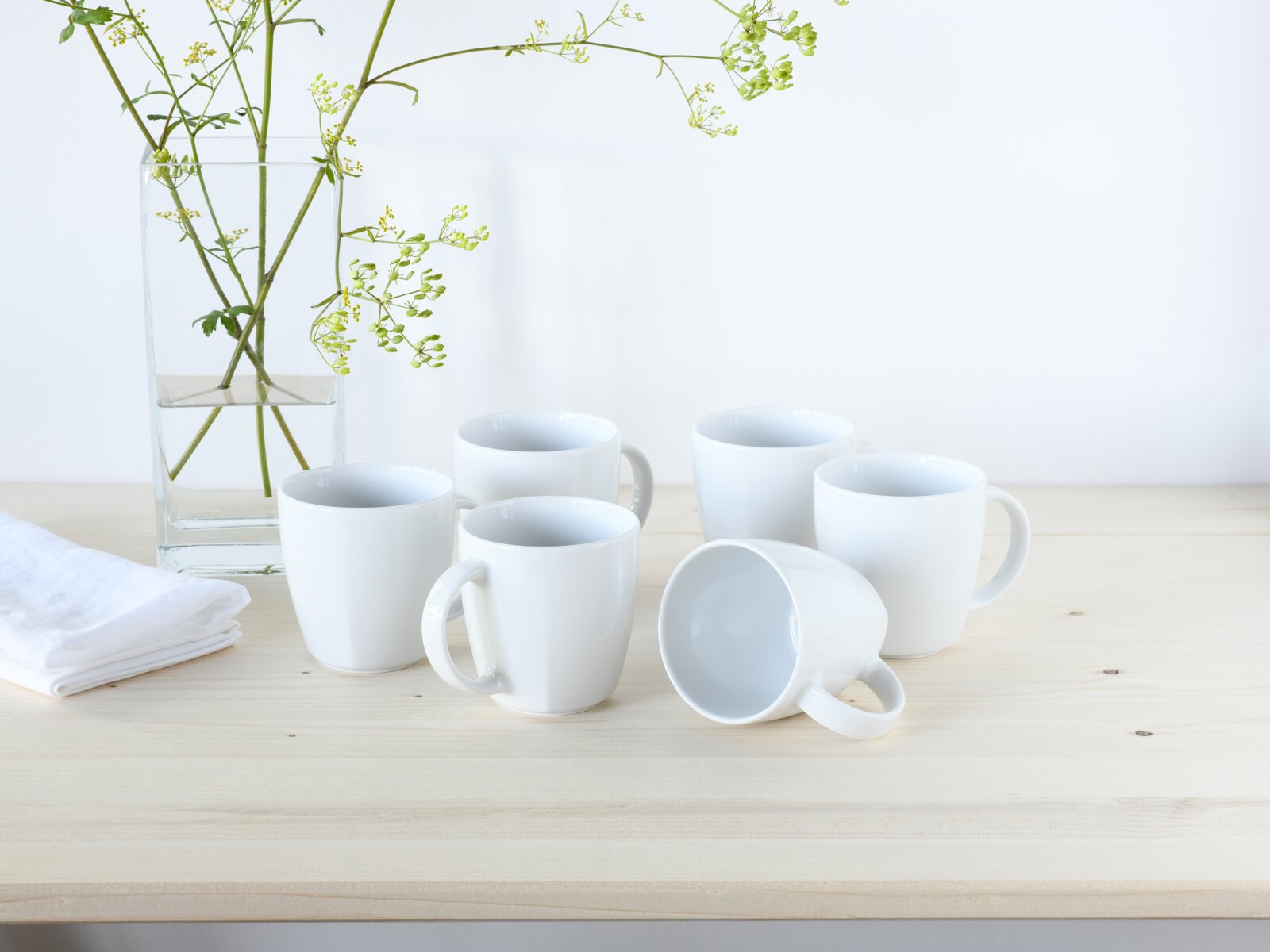 creaTable Kaffeebecher 6-tlg VICTORIA Fünf weiße Keramik-Kaffeetassen auf hellem Holztisch, eine Tasse liegt seitlich, daneben Glasvase mit grünen Zweigen – Aufnahme aus leicht erhöhter Frontperspektive