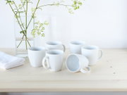 Fünf weiße Keramik-Kaffeetassen auf hellem Holztisch, eine Tasse liegt seitlich, daneben Glasvase mit grünen Zweigen – Aufnahme aus leicht erhöhter Frontperspektive