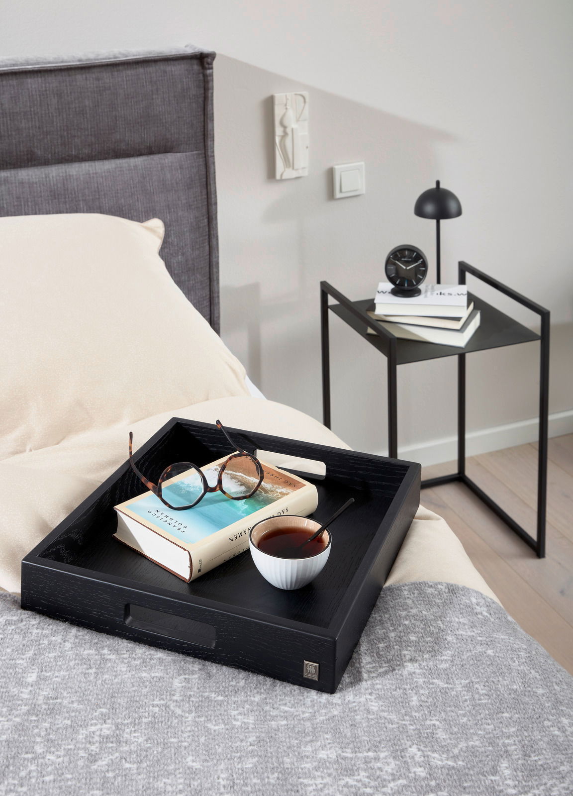 Moderner Beistelltisch in Schwarz neben einem Bett, mit Büchern und einer kleinen Lampe darauf, aus seitlicher Perspektive.