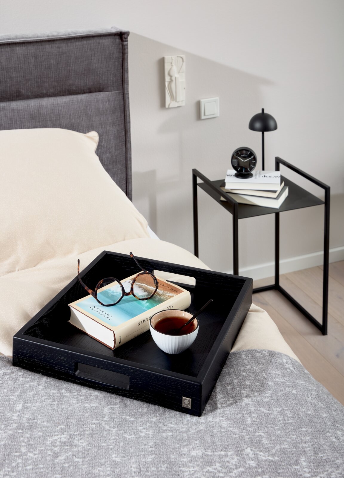 Moderne bijzettafel in het zwart naast een bed, met boeken en een lampje erop, gezien vanuit de zijkant.