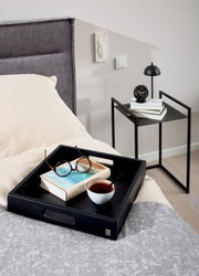 Moderne bijzettafel in het zwart naast een bed, met boeken en een lampje erop, gezien vanuit de zijkant.
