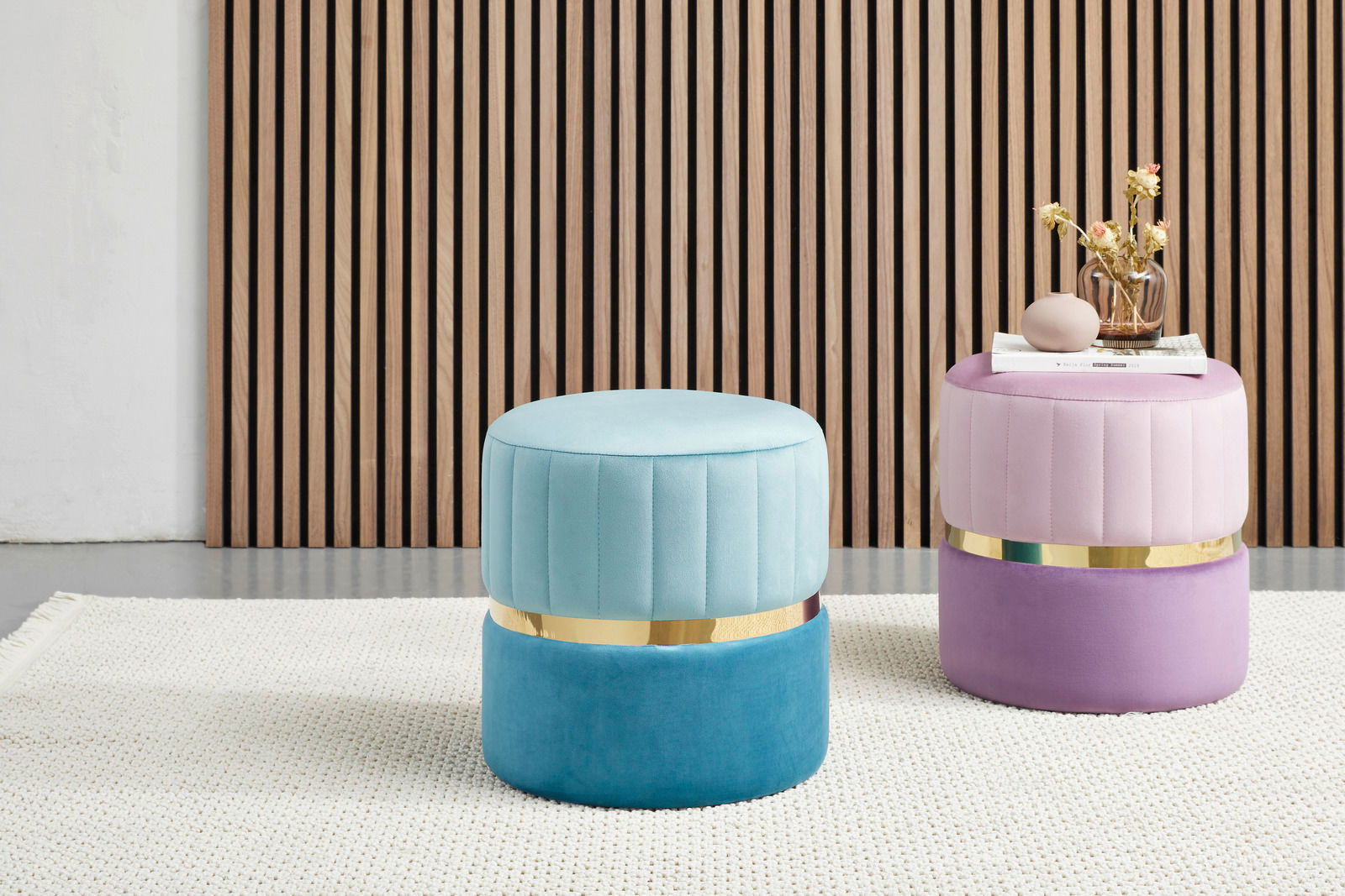 Zwei runde, gepolsterte Hocker in Pastellfarben, einer in Türkis und der andere in Rosa, mit goldenen Akzenten, auf einem hellen Teppich vor einer Holzlamellenwand. Perspektive: Frontalansicht.