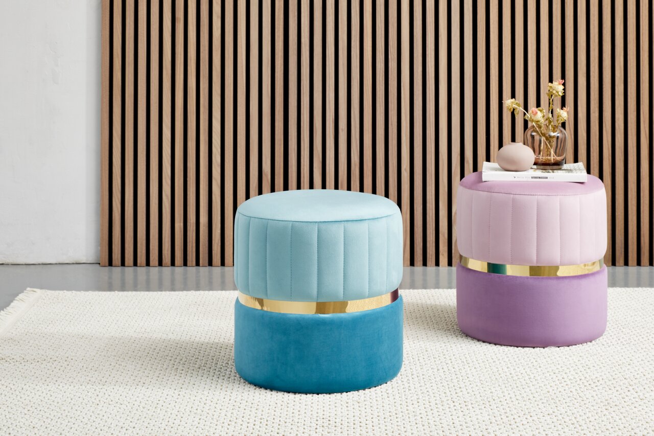 Zwei runde, gepolsterte Hocker in Pastellfarben, einer in Türkis und der andere in Rosa, mit goldenen Akzenten, auf einem hellen Teppich vor einer Holzlamellenwand. Perspektive: Frontalansicht.