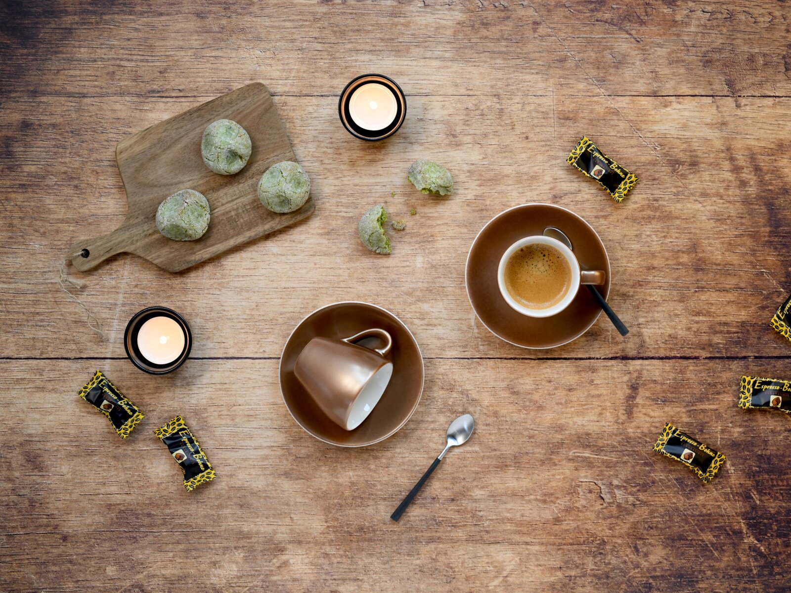 Draufsicht auf rustikalem Holztisch: braune Espressotasse mit Espresso auf Untertasse, zweite Tasse umgedreht, grünes Gebäck auf Holzbrett, Teelichter, Löffel und einzeln verpackte Espresso-Bohnen.