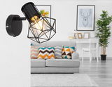 Schwarzer Metallstrahler Globo Lighting PRISKA an einer weißen Wand montiert, seitliche Perspektive, im Hintergrund ein modern eingerichtetes Wohnzimmer mit grauem Sofa und bunten Kissen.