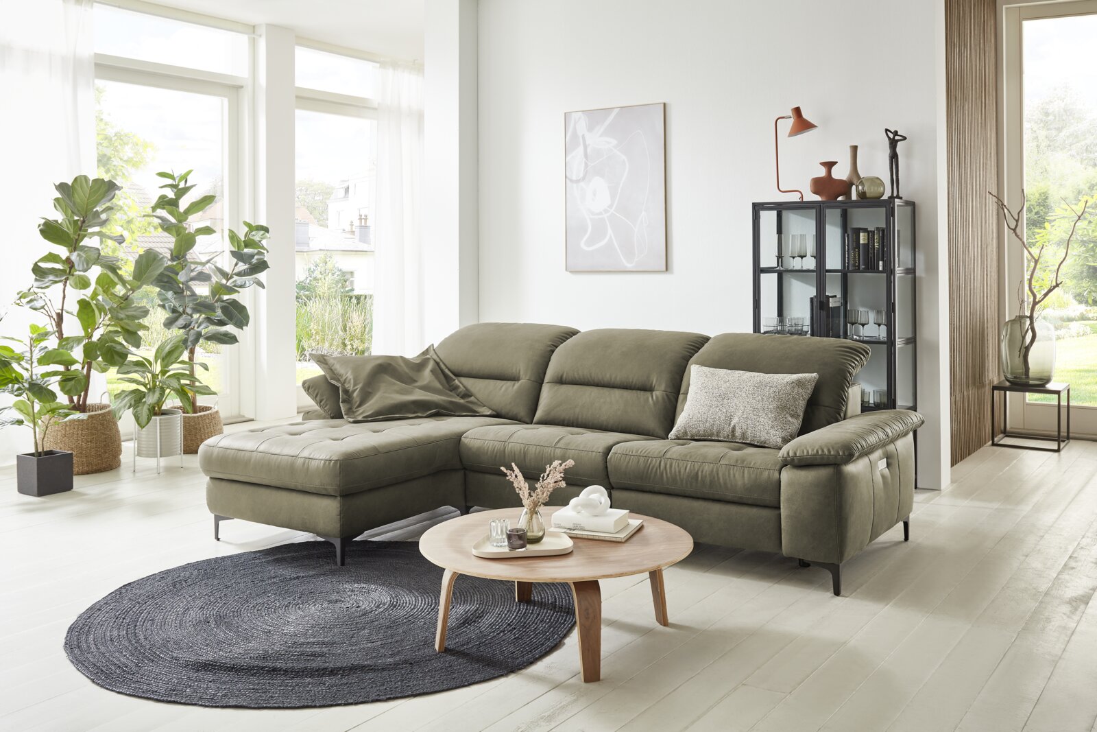 Grüne Longchairkombination in einem modernen Wohnzimmer mit großen Fenstern, Pflanzen und einem runden Holztisch, seitliche Perspektive.