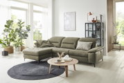 Grüne Longchairkombination in einem modernen Wohnzimmer mit großen Fenstern, Pflanzen und einem runden Holztisch, seitliche Perspektive.