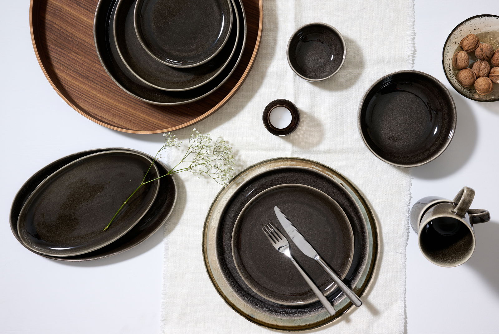 Vogelperspektive auf ein stilvolles Geschirrset in dunklen Erdtönen, bestehend aus Tellern, Schalen und einer Tasse, arrangiert auf einem weißen Tuch mit Besteck und Dekoration.
