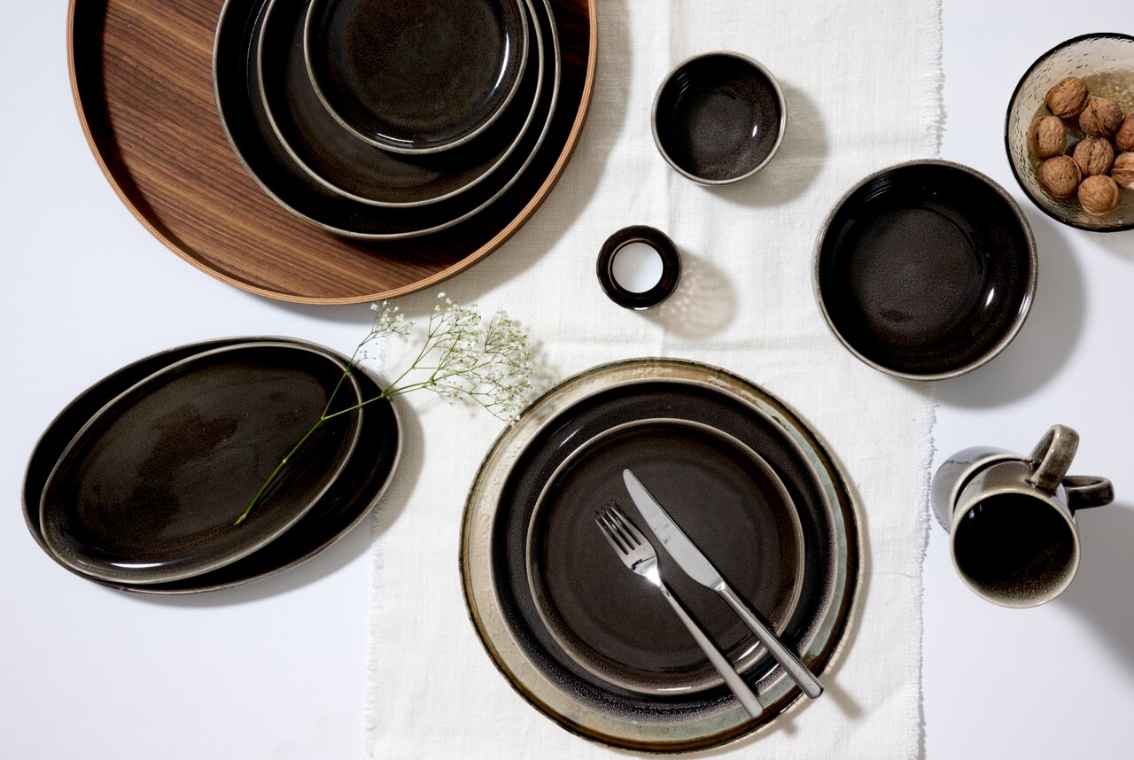 Vogelperspektive auf ein stilvolles Geschirrset in dunklen Erdtönen, bestehend aus Tellern, Schalen und einer Tasse, arrangiert auf einem weißen Tuch mit Besteck und Dekoration.