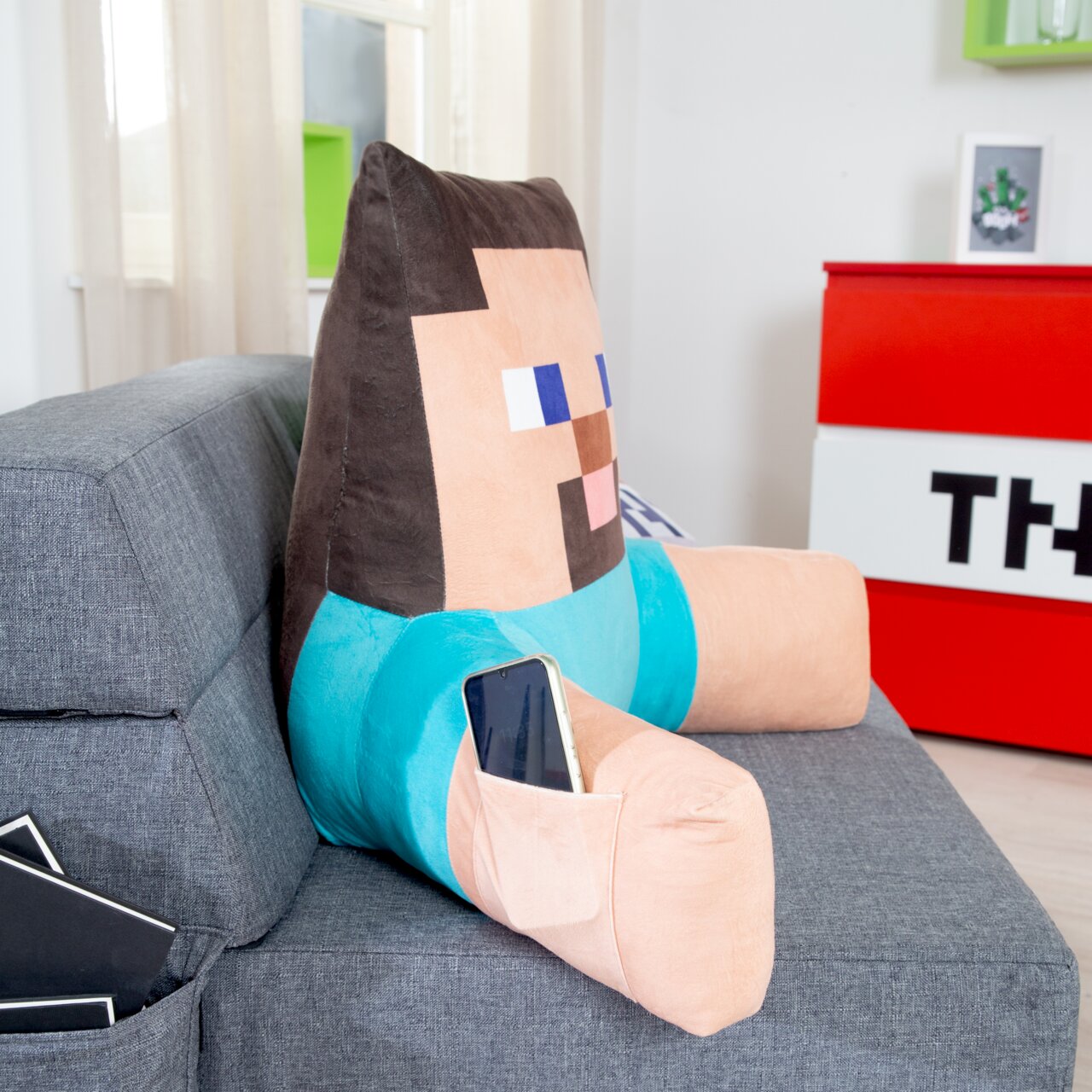 Groot, op Minecraft geïnspireerd Steve-knuffelkussen met een ingebouwd vakje voor een smartphone op de arm, liggend op een grijze bank, gezien vanuit een zijaanzicht