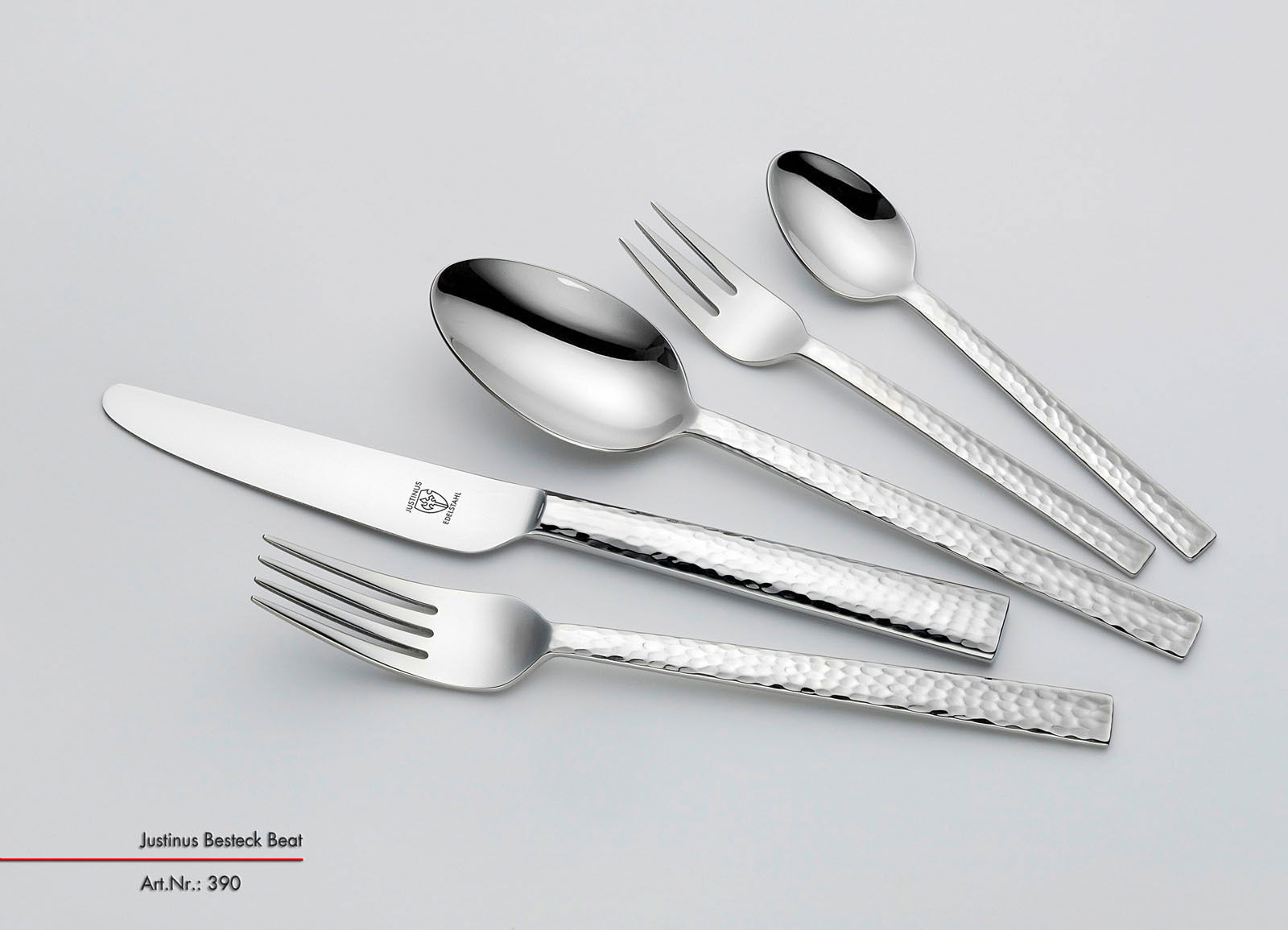 Set aus glänzendem Besteck mit gehämmerten Griffen, bestehend aus Messer, Gabel, Löffel und Teelöffel, in schräger Draufsicht.