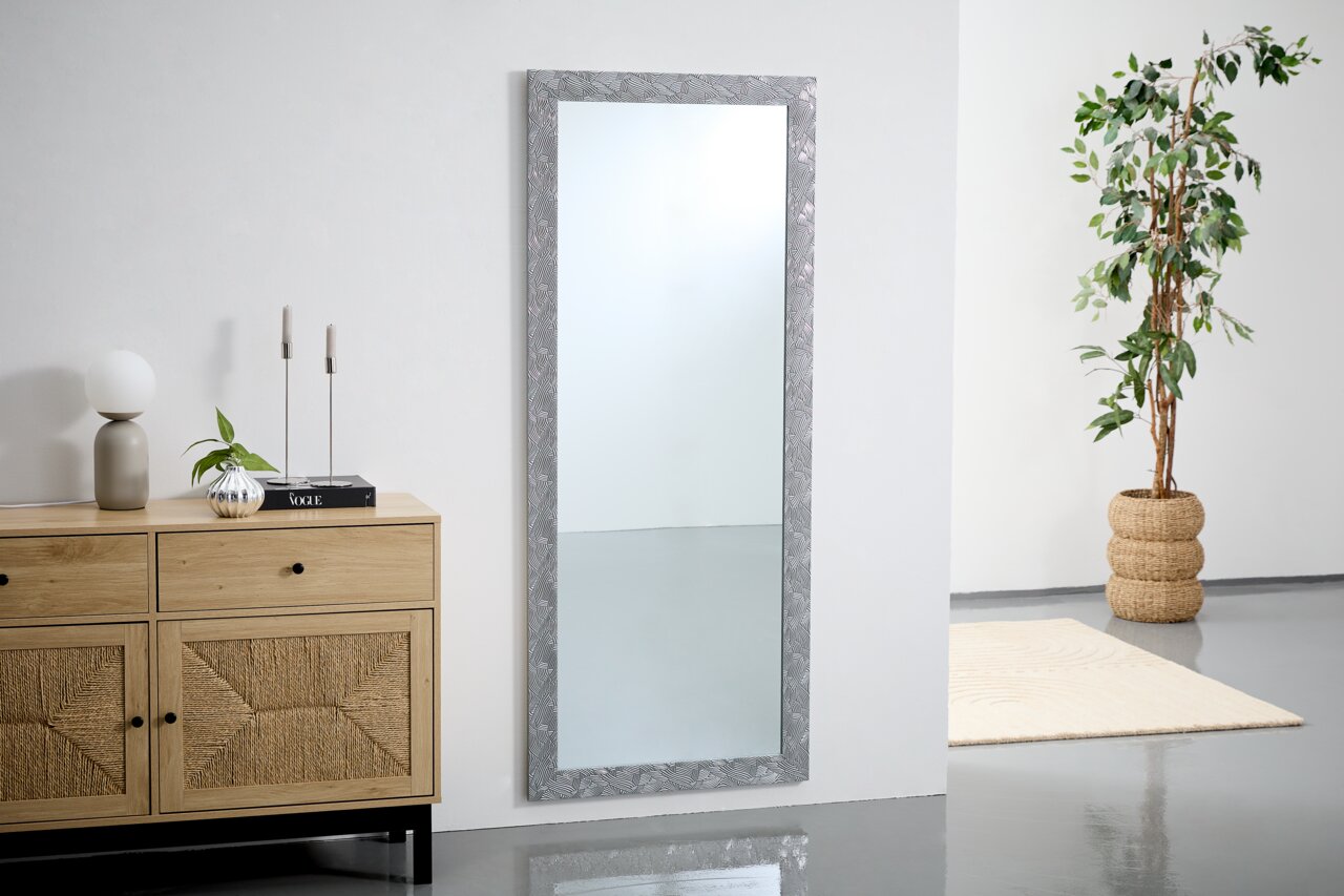 Großer rechteckiger Spiegel mit dekorativem Rahmen, stehend an einer Wand in einem modernen Raum mit Pflanzen und Möbeln, seitliche Perspektive.