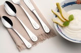 Kinderbesteck-Set aus vier Teilen mit eingravierten Motiven, auf einem beigen Tuch liegend, neben einer Schale mit Joghurt und Birnenscheiben, aus der Vogelperspektive fotografiert.