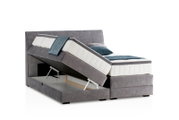 Boxspringbett in seitlicher Perspektive mit geöffnetem Bettkasten und grauem Stoffbezug.