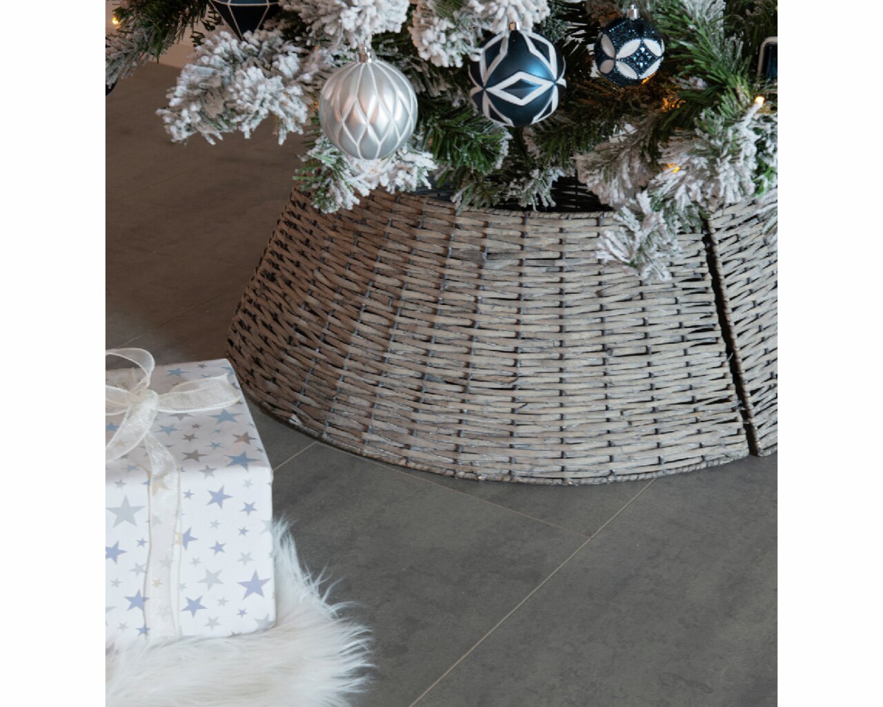 Nahaufnahme eines geflochtenen Baumrings aus Weide, der den unteren Teil eines Weihnachtsbaums umgibt, mit dekorativen Kugeln und einem Geschenk im Vordergrund.