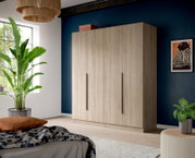 Großer Kleiderschrank in heller Holzoptik mit vier Türen und langen schwarzen Griffen, in einem modernen Schlafzimmer vor dunkelblauer Wand, Perspektive: frontale Ansicht mit leichtem Blick von links.