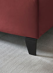 Nahaufnahme eines Boxspringbettbeins mit rotem Stoffbezug und schwarzem Holzfuß auf hellem Teppichboden.