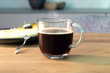 Glas Tasse Napoli 280ml mit heißem Kaffee, seitliche Perspektive, im Hintergrund ein Teller mit Kuchen.