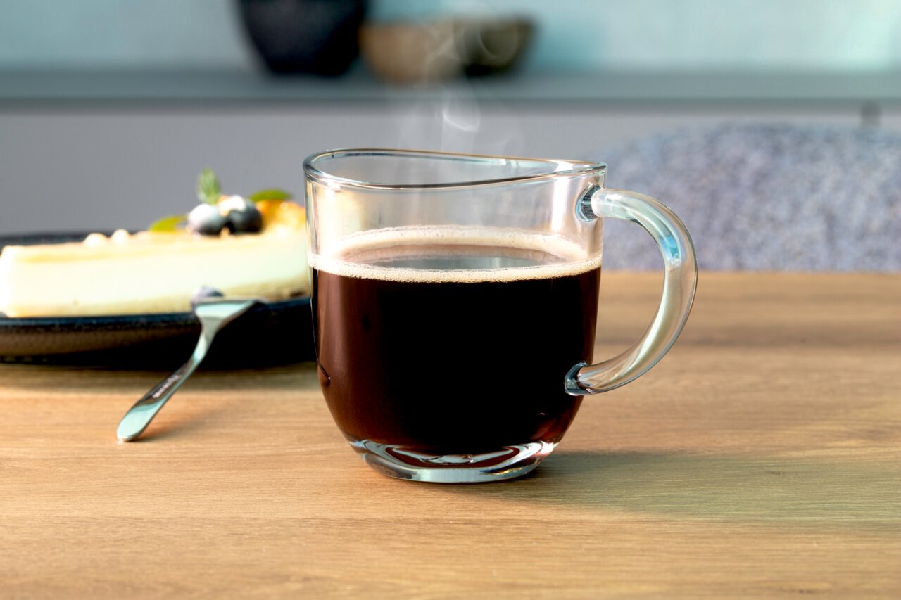 Glas Tasse Napoli 280ml mit heißem Kaffee, seitliche Perspektive, im Hintergrund ein Teller mit Kuchen.