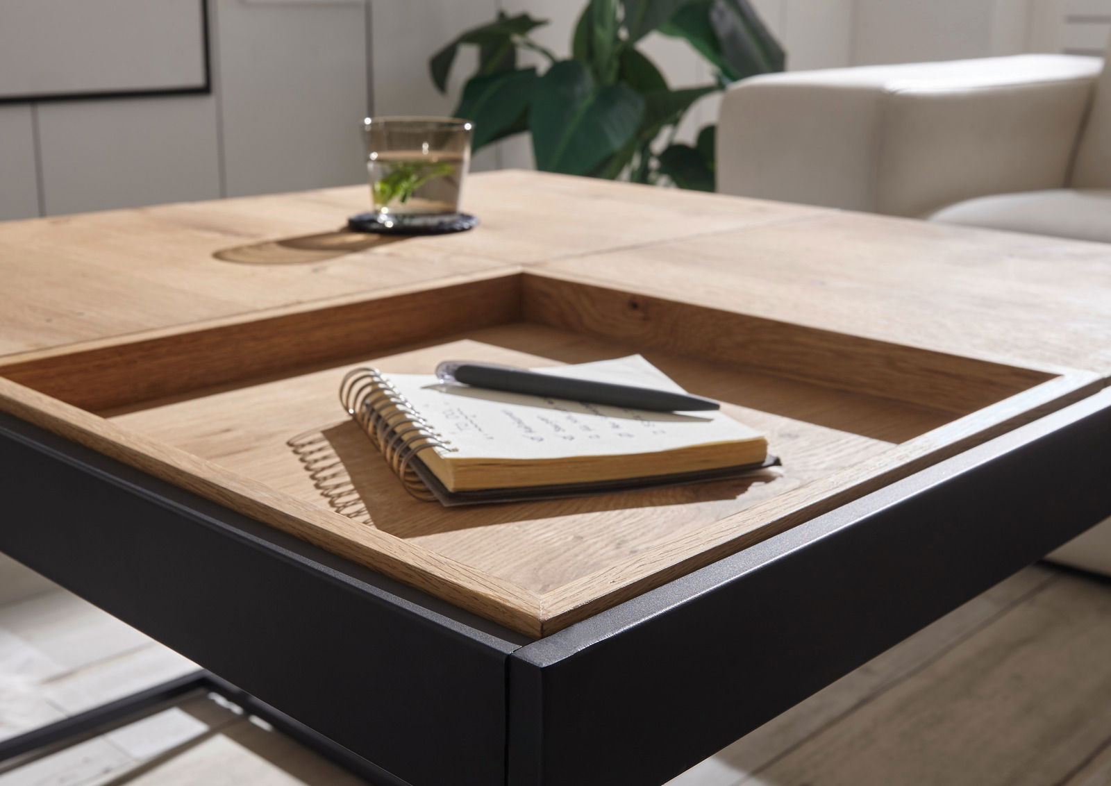 Nahaufnahme eines modernen Couchtisches mit Holzoberfläche und schwarzem Metallrahmen, darauf ein Notizbuch und ein Stift, im Hintergrund eine Pflanze und ein Sofa.