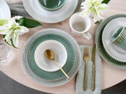 Draufsicht auf ein stilvoll gedecktes Tischarrangement mit grün-grauen Keramiktellern, weißer Schale mit goldenem Löffel, Tasse, goldfarbenem Besteck auf Stoffserviette und weißen Lilien auf hellem Holztisch.