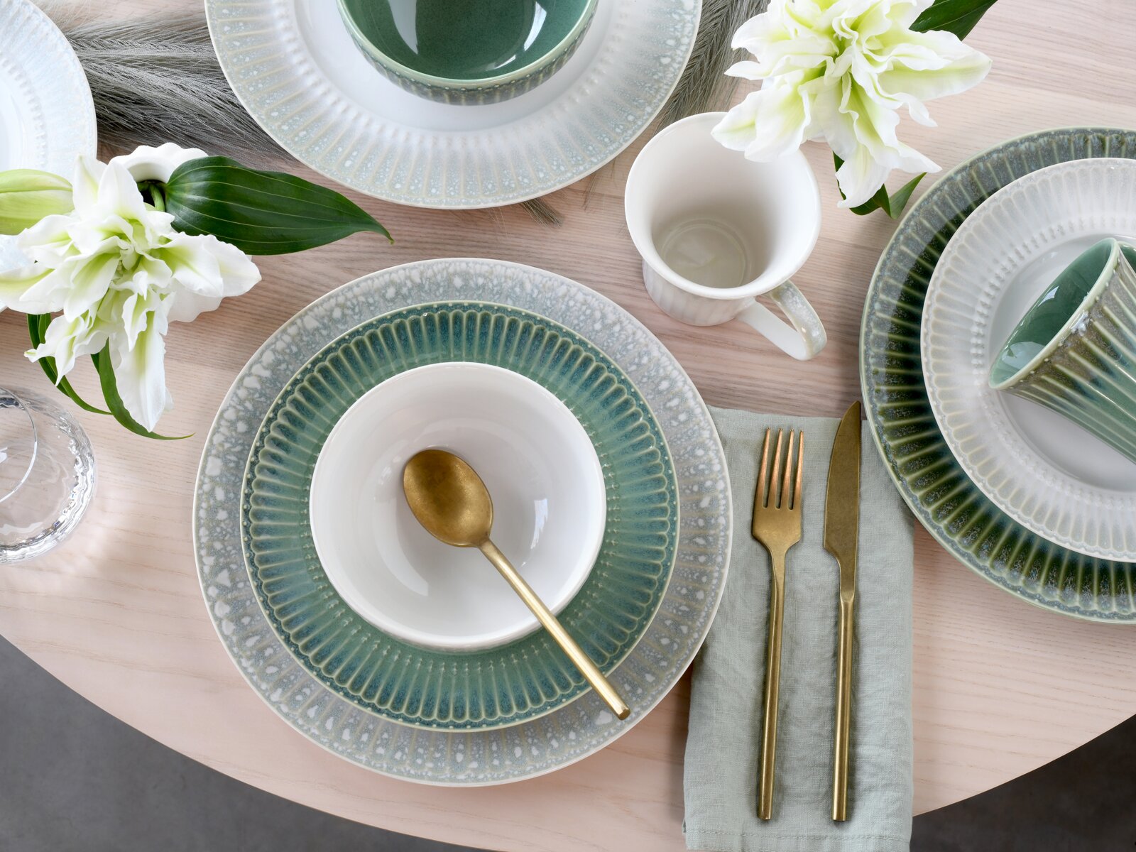 Draufsicht auf ein stilvoll gedecktes Tischarrangement mit grün-grauen Keramiktellern, weißer Schale mit goldenem Löffel, Tasse, goldfarbenem Besteck auf Stoffserviette und weißen Lilien auf hellem Holztisch.