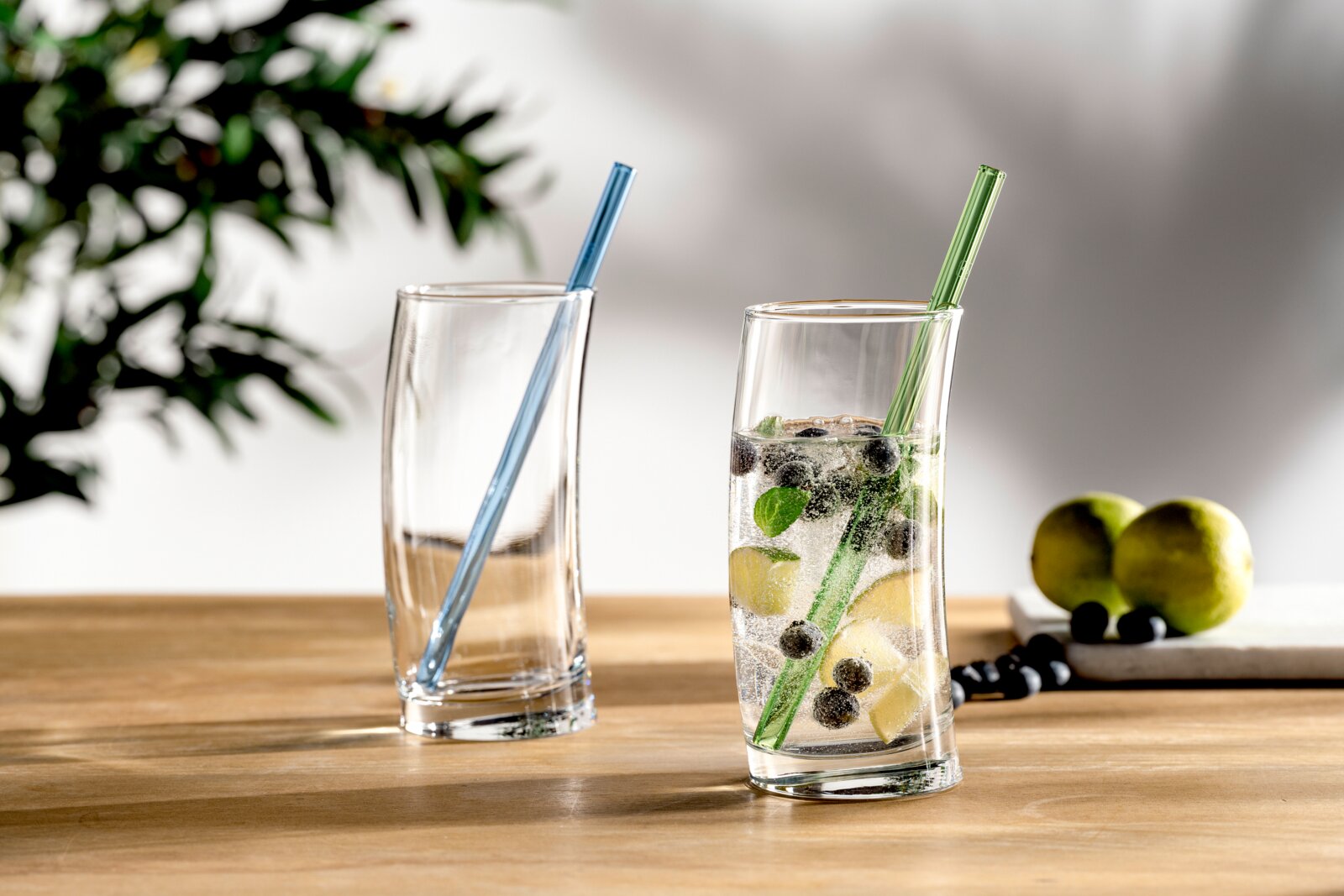 Zwei transparente Trinkgläser auf Holztisch, eines mit blauem und eines mit grünem Glasstrohhalm im rechten Glas sprudelndes Getränk mit Eis, Limettenstücken, Blaubeeren und Minze, Perspektive: leicht seitliche Frontansicht auf Tischhöhe.