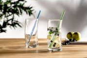 Zwei transparente Trinkgläser auf Holztisch, eines mit blauem und eines mit grünem Glasstrohhalm; im rechten Glas sprudelndes Getränk mit Eis, Limettenstücken, Blaubeeren und Minze, Perspektive: leicht seitliche Frontansicht auf Tischhöhe.