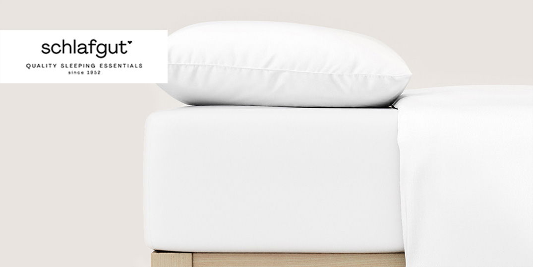 Nahaufnahme eines weißen Bettes mit einem Kissen mit dem Schlafgut-Logo darüber.