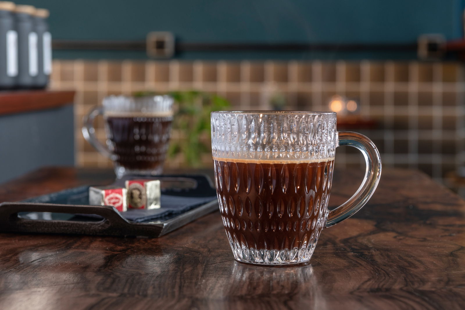Nahaufnahme eines Barista Kaffeebechers aus Glas mit strukturiertem Design, gefüllt mit schwarzem Kaffee, auf einem Holztisch in einem gemütlichen Café-Ambiente.