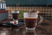 Nahaufnahme eines Barista Kaffeebechers aus Glas mit strukturiertem Design, gefüllt mit schwarzem Kaffee, auf einem Holztisch in einem gemütlichen Café-Ambiente.