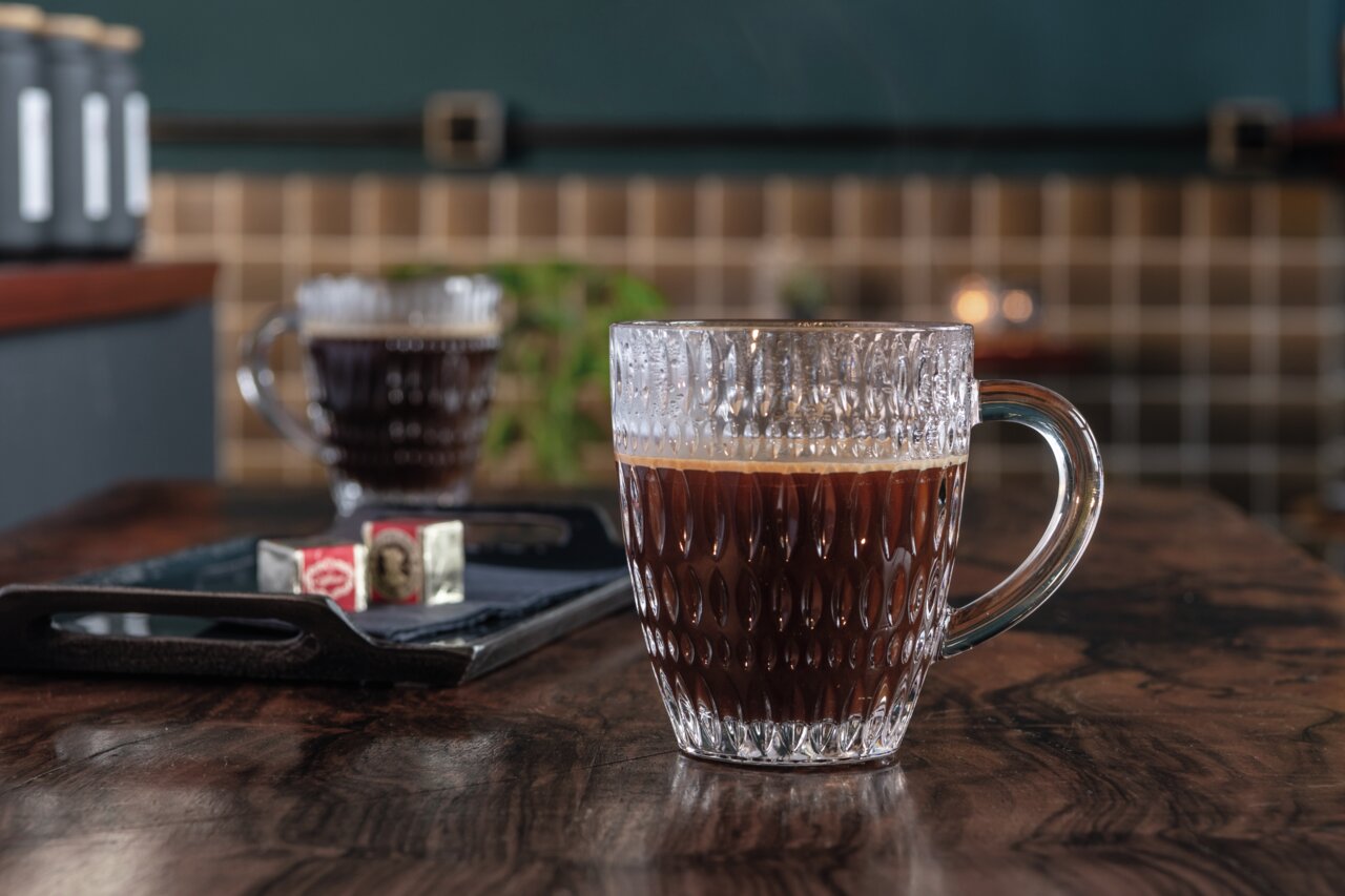 Nahaufnahme eines Barista Kaffeebechers aus Glas mit strukturiertem Design, gefüllt mit schwarzem Kaffee, auf einem Holztisch in einem gemütlichen Café-Ambiente.